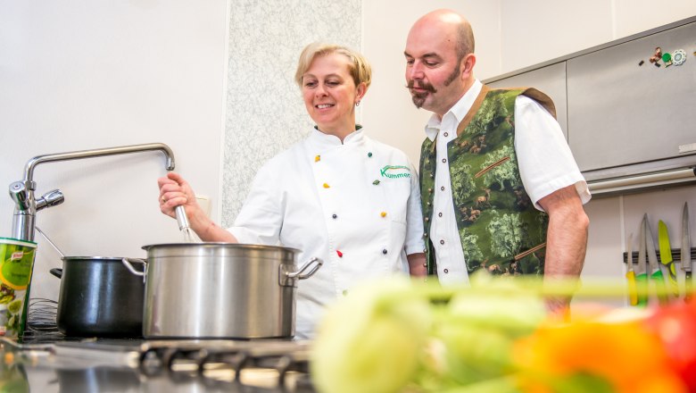 Zwei Personen in einer Küche, eine rührt in einem Topf, die andere schaut zu.