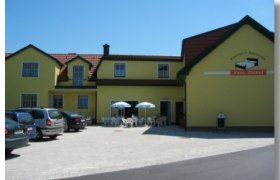 Gelbes Gebäude mit der Aufschrift 'Pension Ziekel', Autos und Tische mit Sonnenschirmen davor.