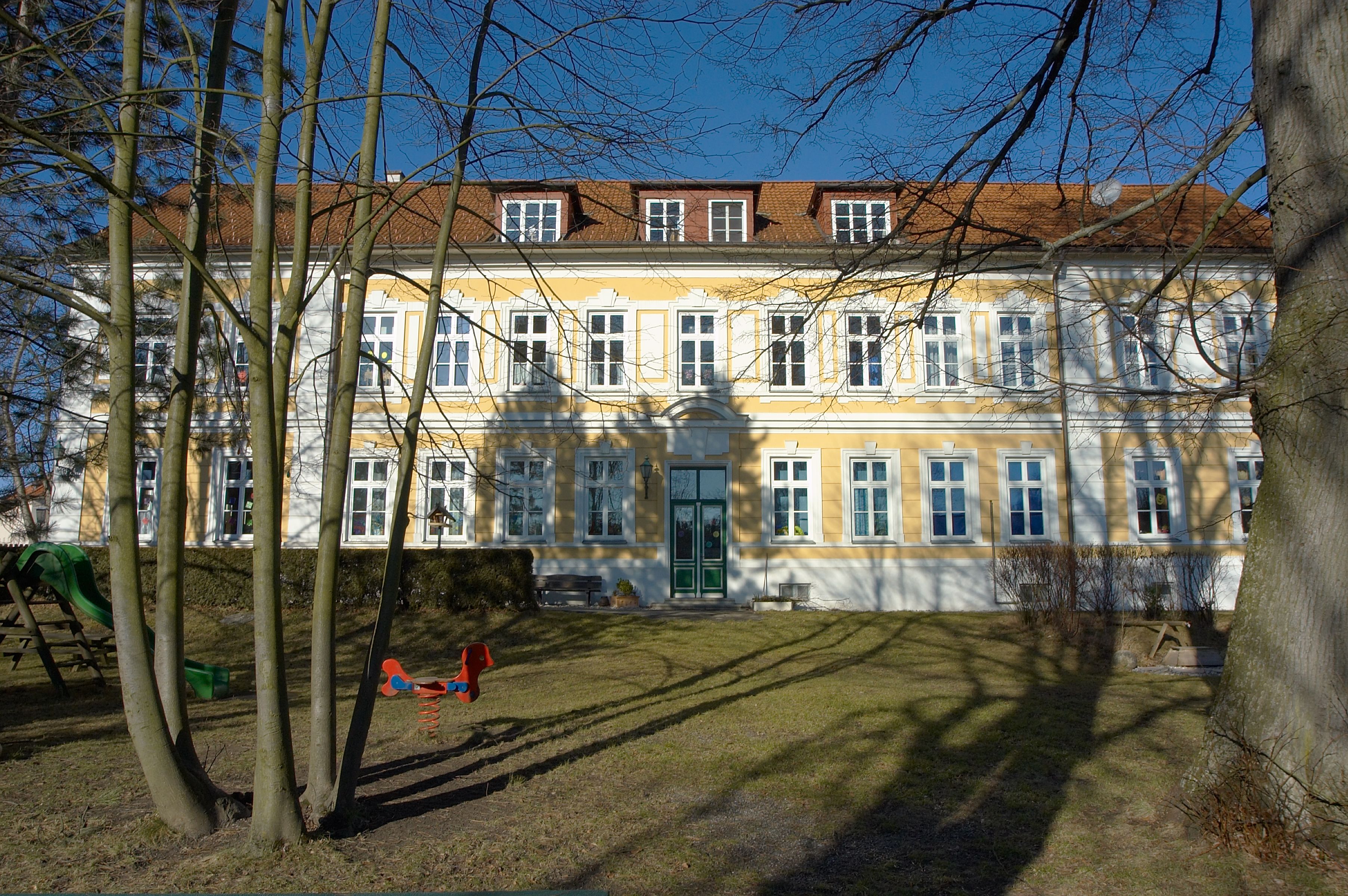 Gelbes Gebäude mit weißen Fensterrahmen und rotem Dach, umgeben von Bäumen und Spielplatz.