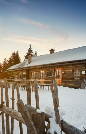 Inmitten einer winterlichen Landschaft erhebt sich die charmante Hütte, umgeben von schneebedeckten Bäumen und sanften Hügeln. Der Sonnenuntergang taucht die Szene in warmes Licht und schafft eine einladende Atmosphäre für Wanderer und Naturliebhaber. Hier, wo die Ruhe der Natur auf die Gemütlichkeit der Almhütte trifft, wird jeder Besuch zu einem unvergesslichen Erlebnis.