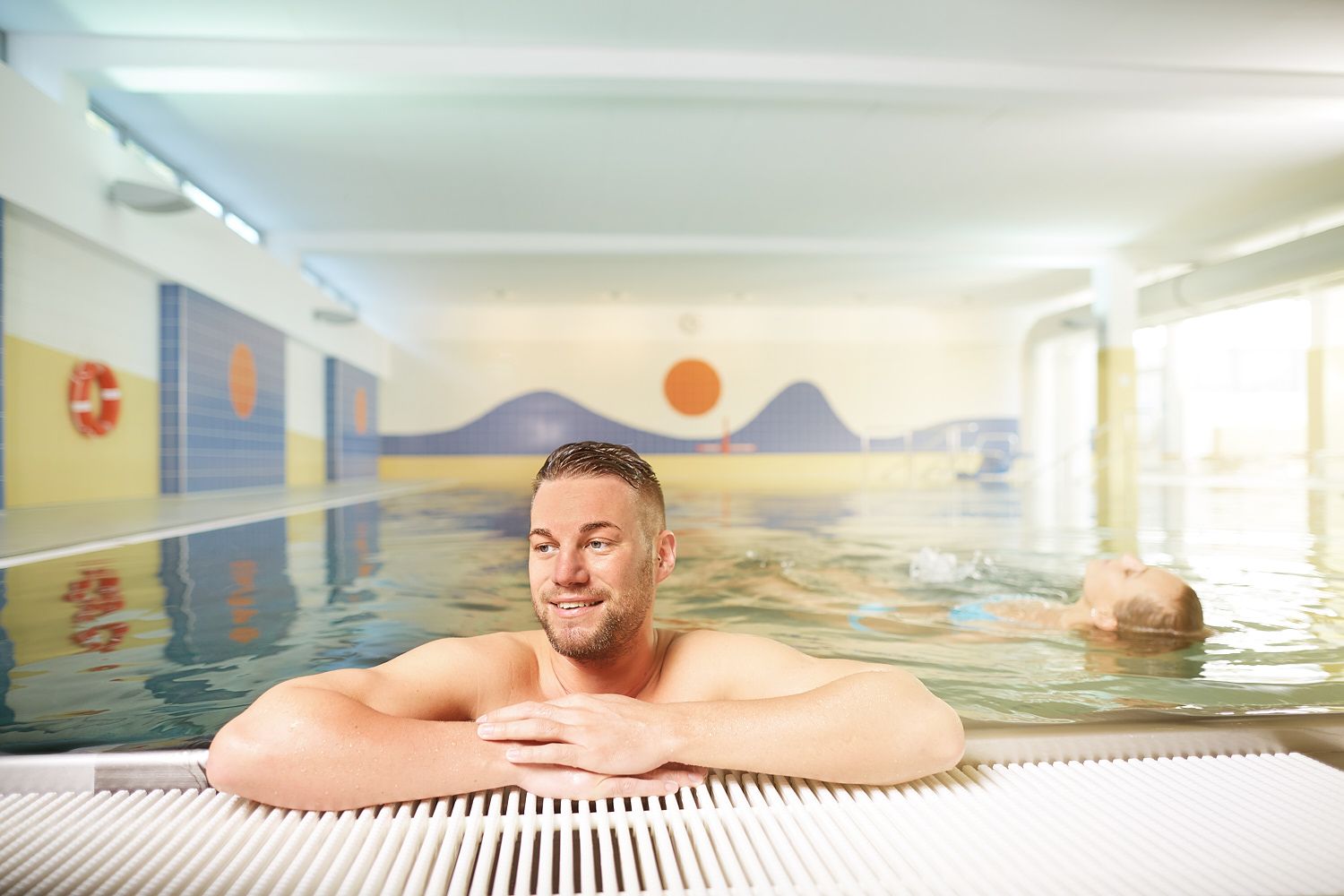 Ein Mann lehnt sich am Beckenrand eines Hallenbads an, während im Hintergrund eine Person im Wasser schwimmt.