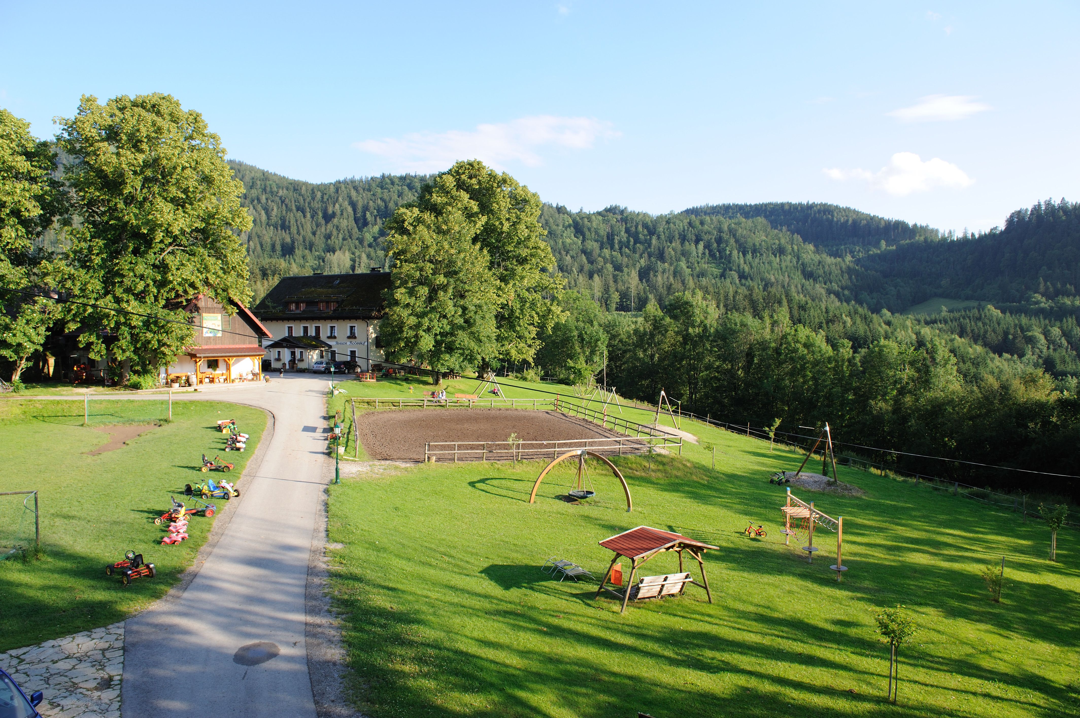 Ein Bauernhof in einer grünen, hügeligen Landschaft mit Spielplatz und Kettcars auf einer Wiese.