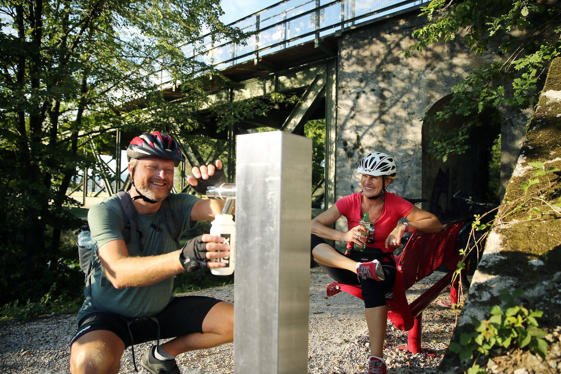 Ein aktives Paar genießt eine wohlverdiente Pause am Trinkbrunnen, umgeben von der malerischen Natur des Erlauftals. Die sanften Hügel und das plätschernde Wasser schaffen eine entspannende Atmosphäre, die zum Verweilen einlädt.