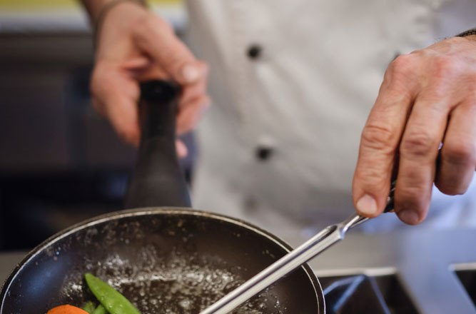 A cook in a white uniform fries vegetables in a pan on a gas stove.