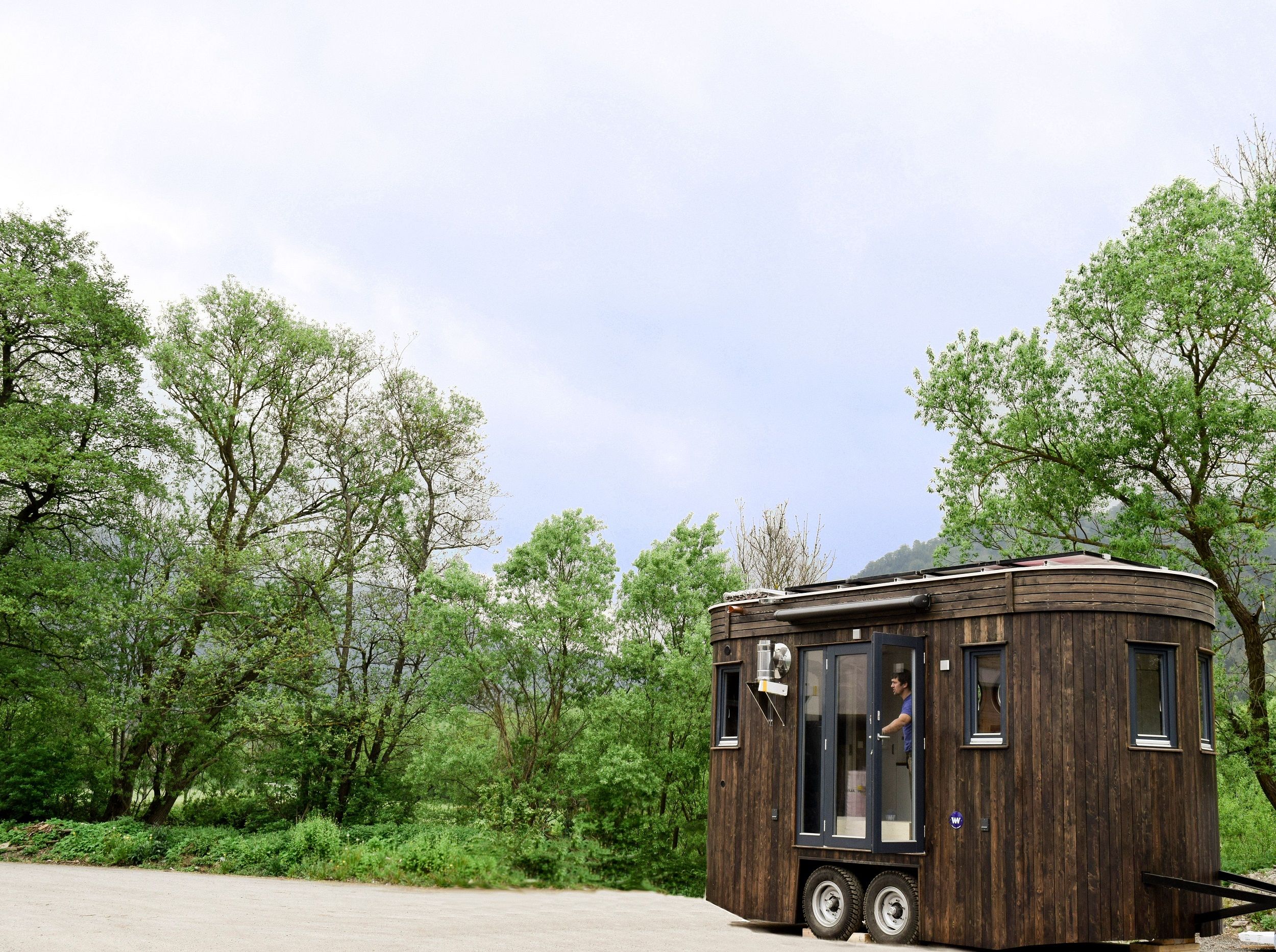 Ein kleiner, dunkler Wohnwagen mit Holzverkleidung steht auf einem Parkplatz vor grünen Bäumen und bewölktem Himmel.