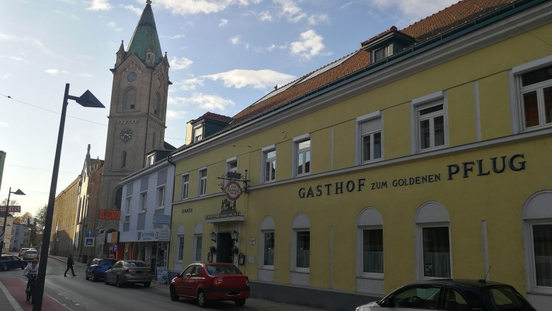 Gasthof zum goldenen Pflug, © Roman Zöchlinger Straßenszene mit gelbem Gebäude 'Gasthof zum goldenen Pflug' und Kirchturm im Hintergrund.