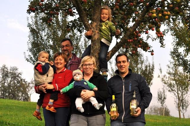 A family is standing in front of an apple tree in a meadow. Two adults are holding small children, one child is sitting in the tree. A man is holding two bottles.