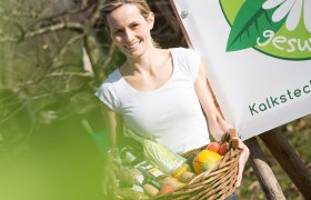 Frau mit Korb voller Gemüse vor einem Schild mit der Aufschrift 'gesund'.