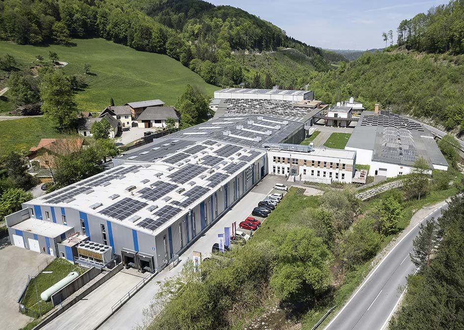 Luftaufnahme einer großen Fabrikanlage mit Solarpanelen auf den Dächern, umgeben von grüner Landschaft.
