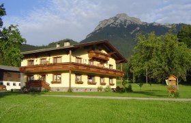 Ein traditionelles Bauernhaus vor einem Berg in einer gr&uuml;nen Landschaft.