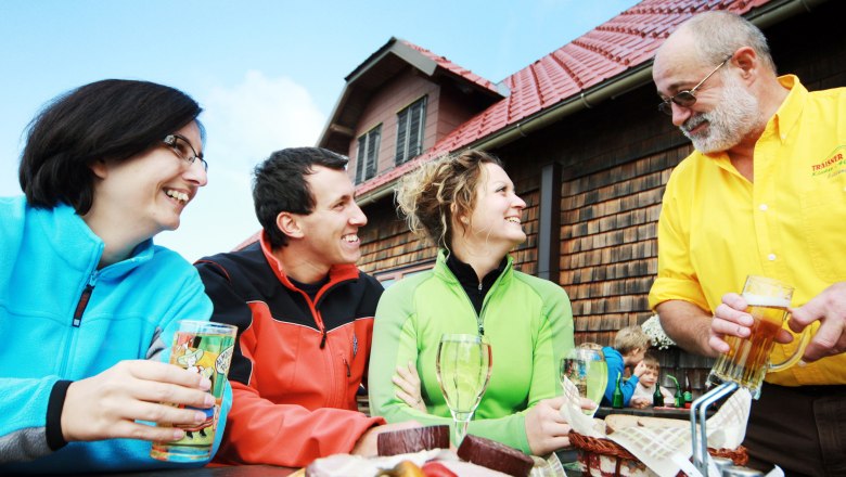 Traisner Huts, © weinfranz.at Three people sit laughing at a table in front of a hut while a man in a yellow shirt serves drinks.