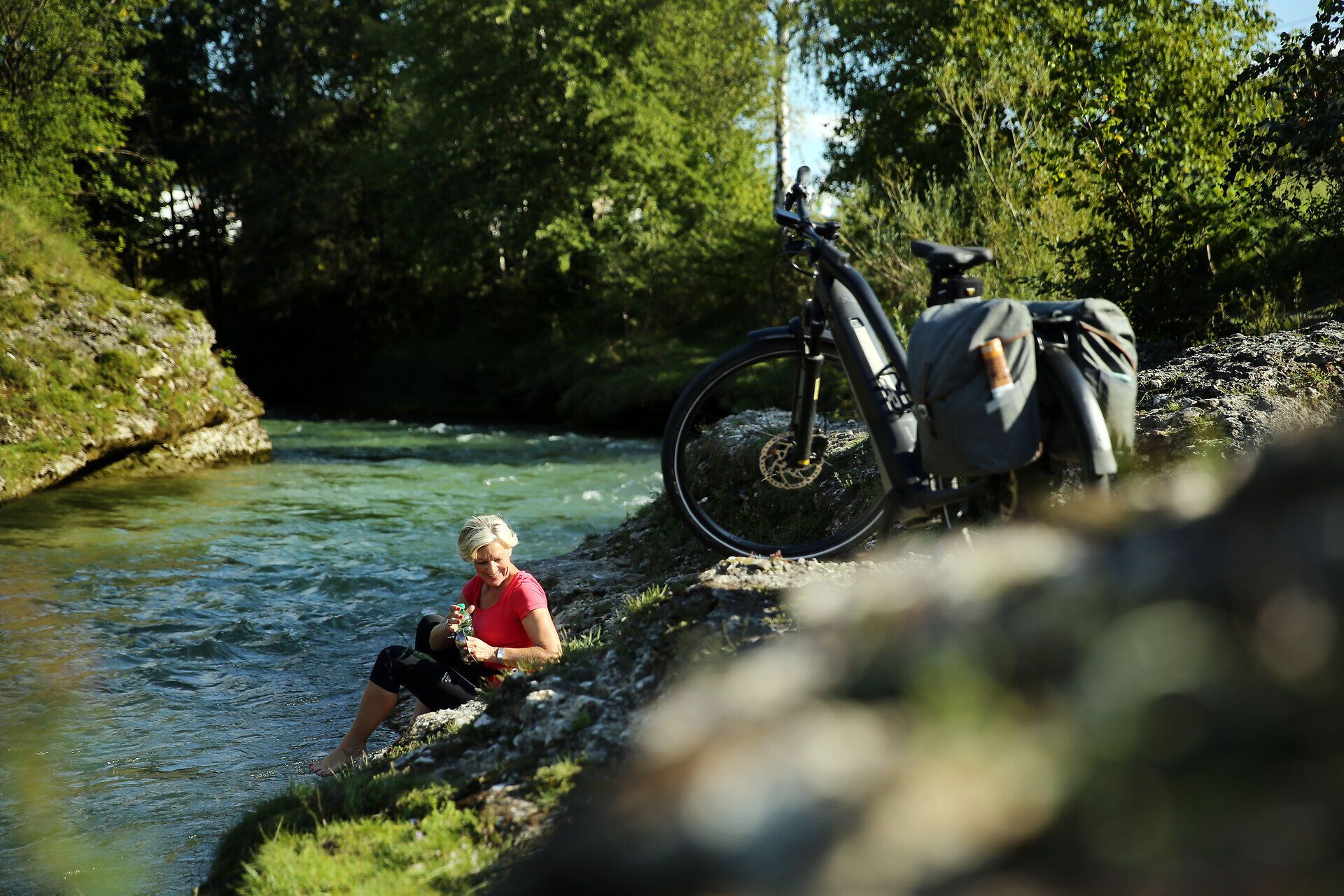 Ein Radfahrer genießt eine wohlverdiente Pause am Ufer eines glitzernden Flusses, umgeben von üppigem Grün und der sanften Melodie der Natur. Die erfrischende Brise und das sanfte Plätschern des Wassers laden dazu ein, die Seele baumeln zu lassen und die Schönheit der Umgebung zu erleben.