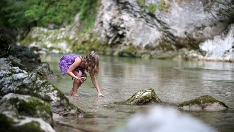 A child in a purple dress plays in a clear stream, surrounded by rocks and green nature.