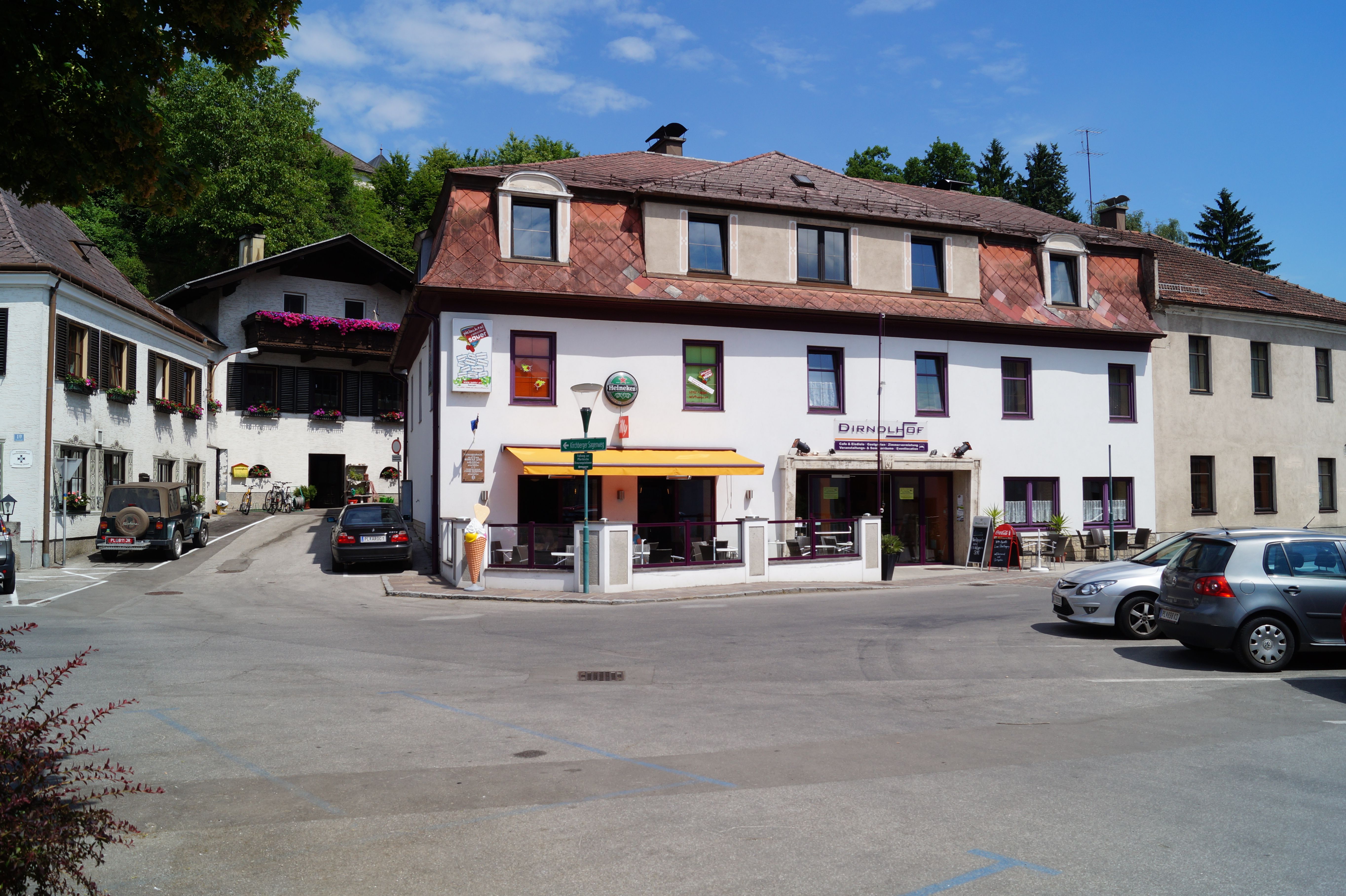 Ein Gebäude mit der Aufschrift 'Dirndlhof' und einer Terrasse mit Sonnenschirm. Autos sind auf dem Parkplatz davor geparkt.