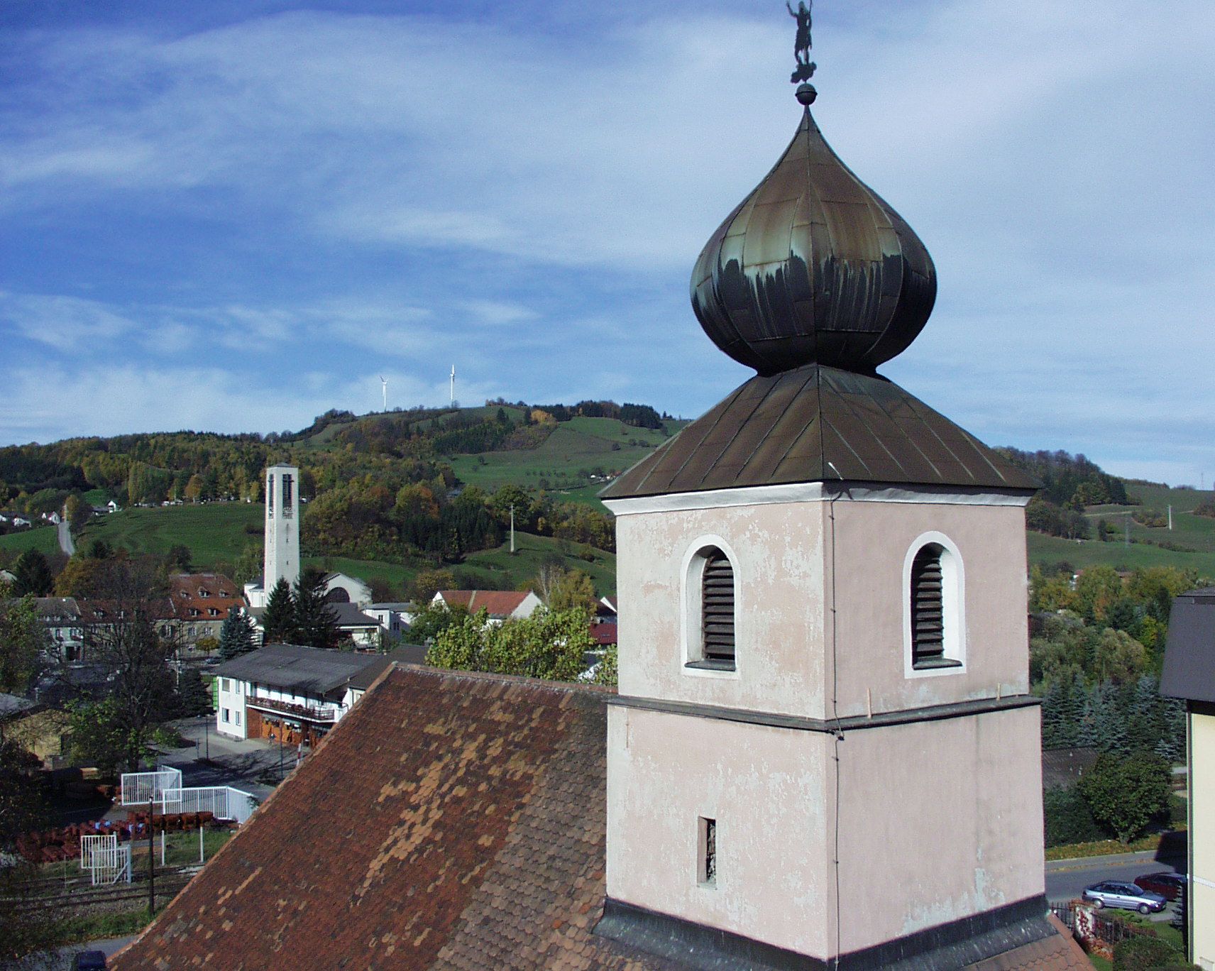 Kirchturm mit Zwiebeldach vor hügeliger Landschaft und Windrädern.