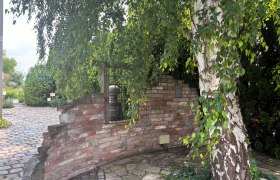 Ein gepflasterter Weg führt an einer alten Backsteinmauer und einem großen Baum vorbei in einen grünen Garten.