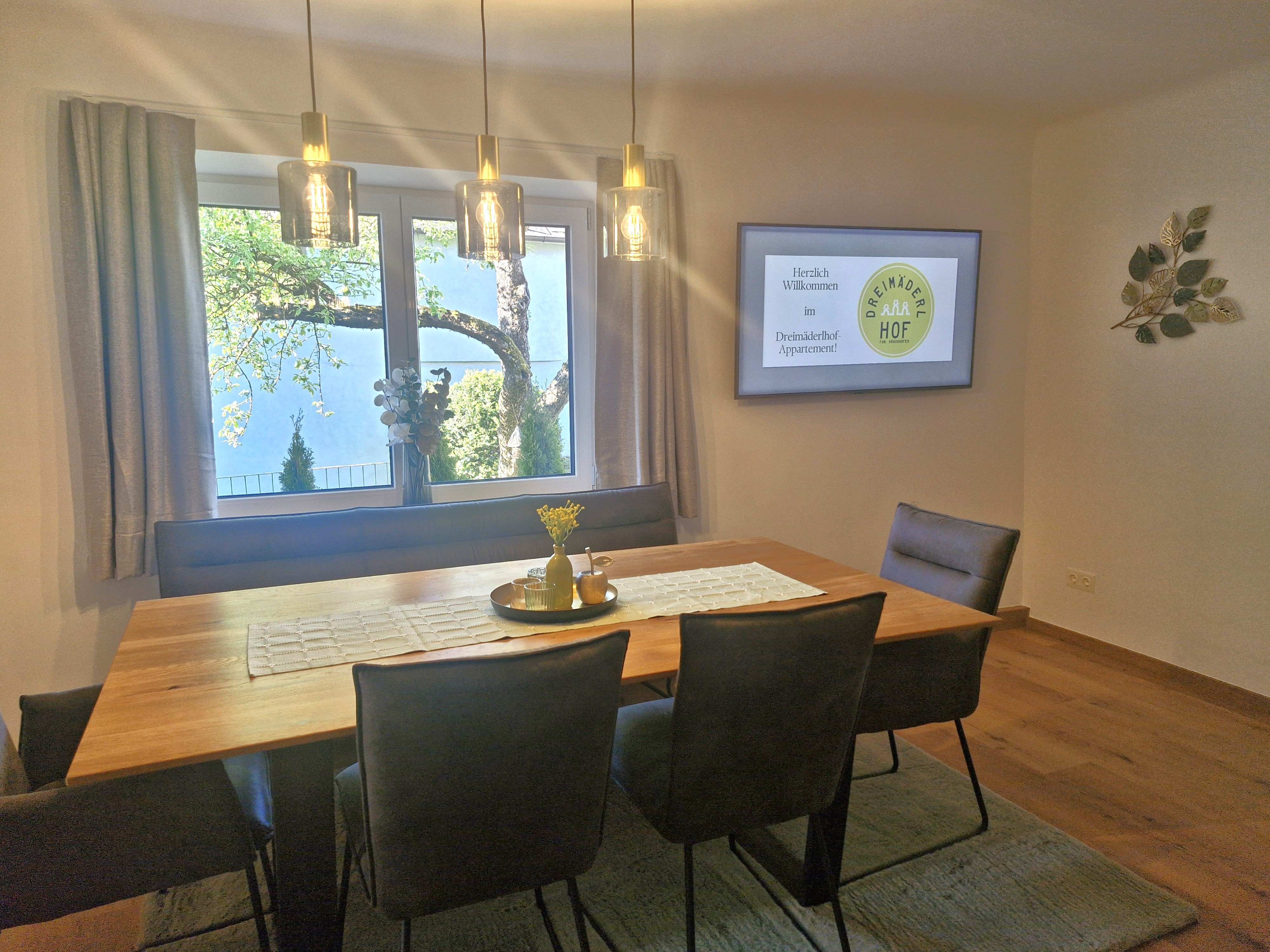 Esszimmer mit Holztisch, Stühlen und Fenster mit Blick auf einen Baum. An der Wand hängt ein Bildschirm mit Begrüßungstext.