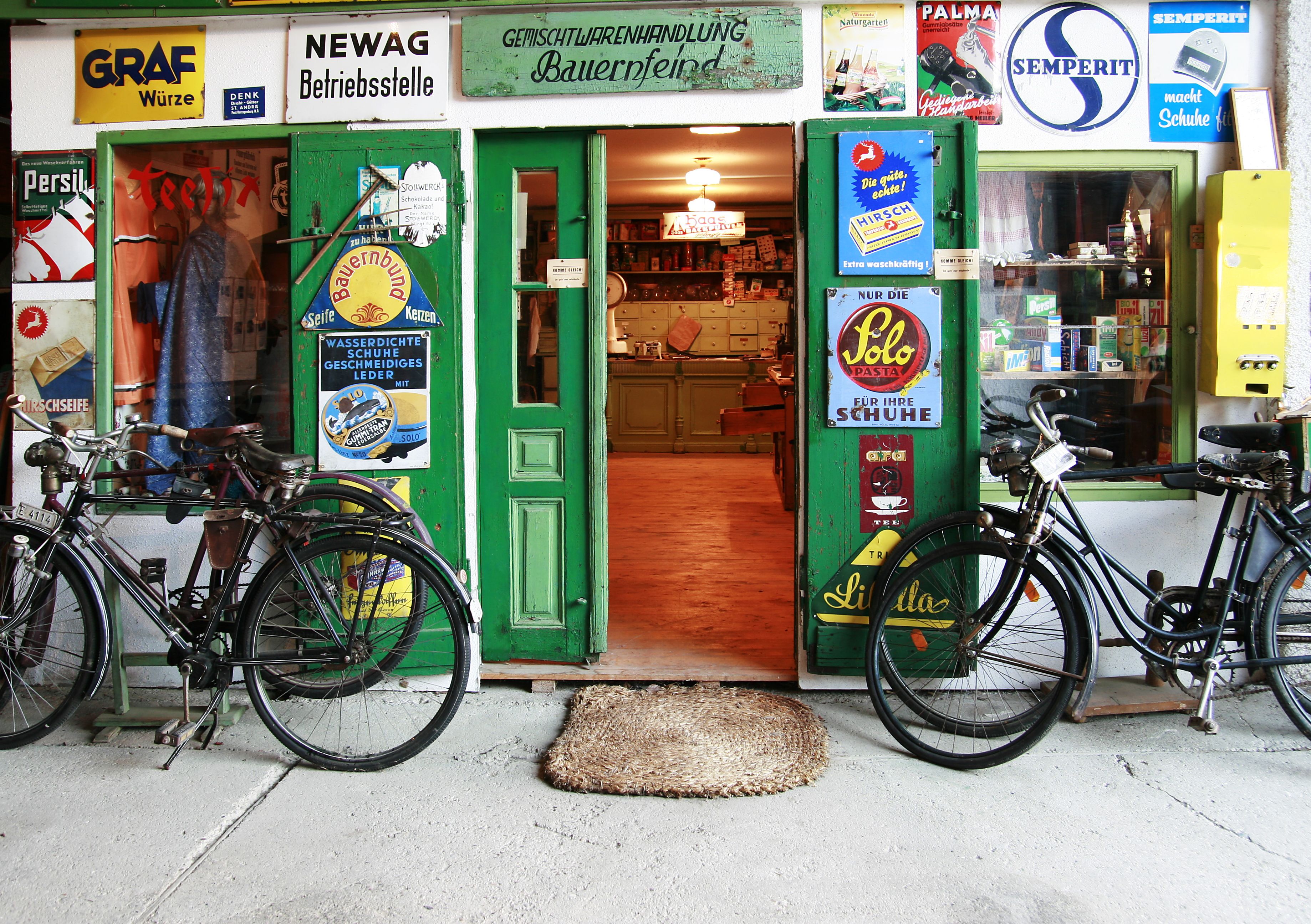 Eingang eines alten Ladens mit grüner Tür, umgeben von Retro-Werbeschildern und zwei Fahrrädern.