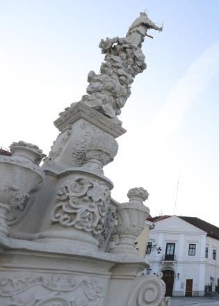 Nahaufnahme einer kunstvoll verzierten S&auml;ule mit Skulpturen und Ornamenten, im Hintergrund Geb&auml;ude und blauer Himmel.