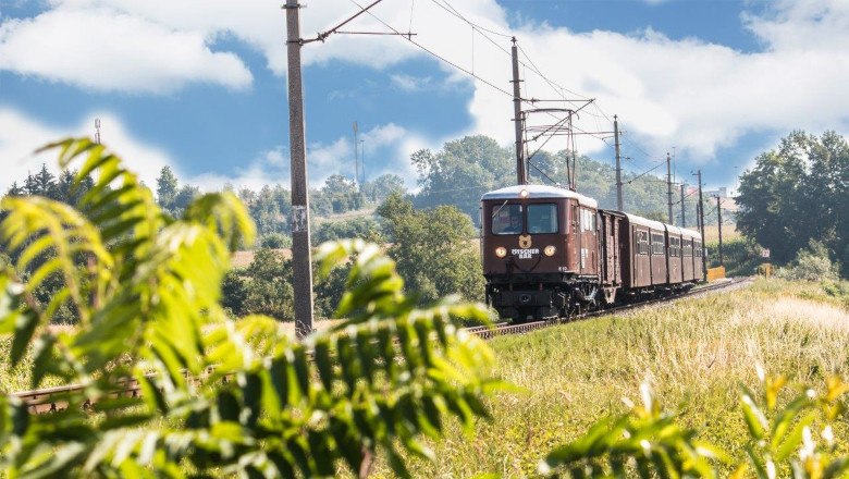 Ein nostalgischer Zug f&auml;hrt durch eine gr&uuml;ne Landschaft mit blauem Himmel.