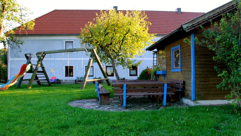 Ein Spielplatz mit Schaukel, Rutsche und Sitzbänken vor einem Haus mit rotem Dach.