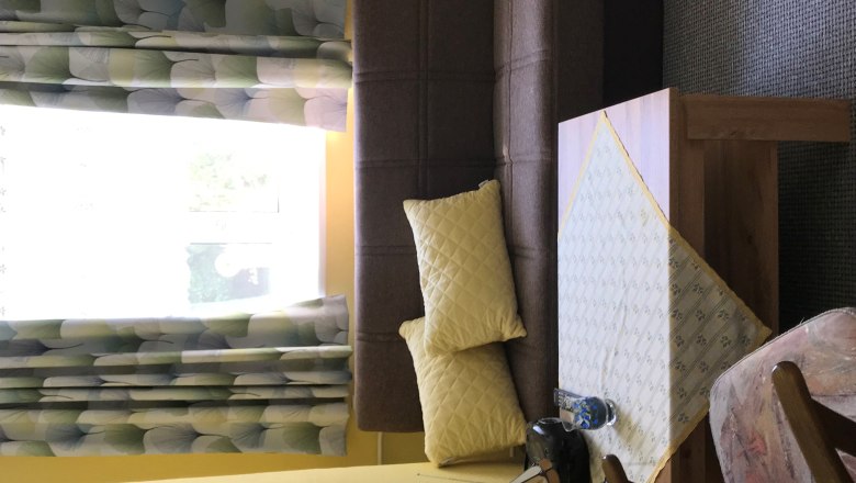 Wohnzimmer mit braunem Sofa, gelben Kissen, Tisch mit Tischdecke, Sessel und Fenster mit gemusterten Vorhängen.