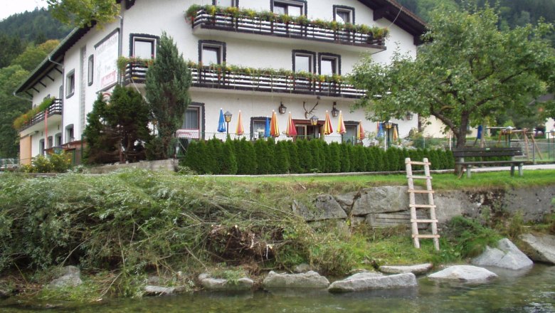 Ein traditionelles Gasthaus mit Balkon und Blumenk&auml;sten, umgeben von B&auml;umen und einem Fluss im Vordergrund.