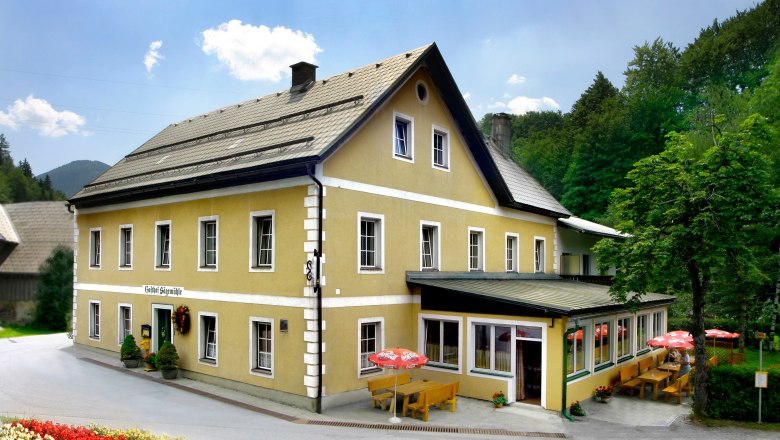 Ein gelbes Gasthaus mit Terrasse und Sonnenschirmen, umgeben von B&auml;umen.
