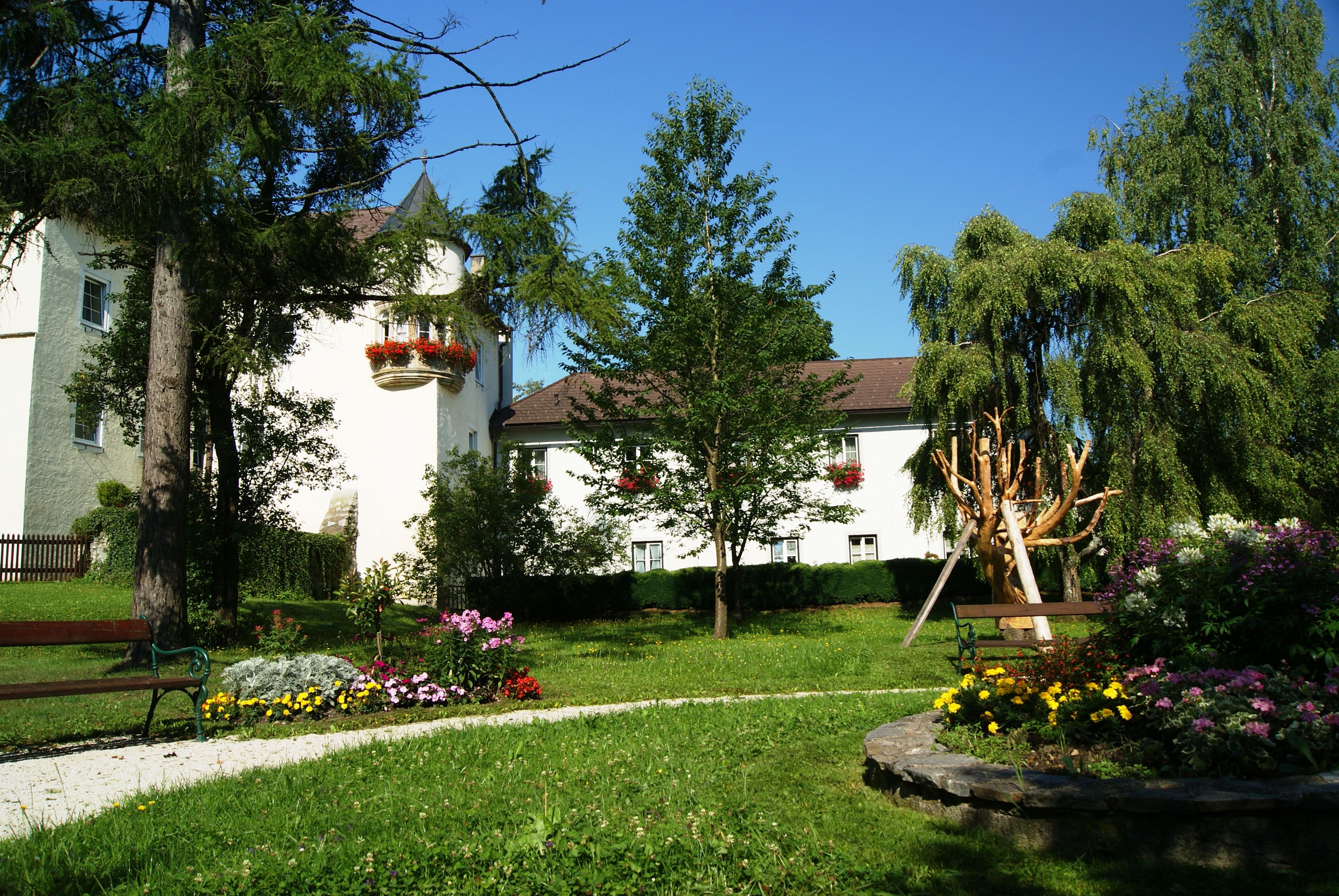 Ein Schloss mit weißer Fassade und roten Blumen, umgeben von einem gepflegten Garten mit Bäumen und Blumenbeeten.