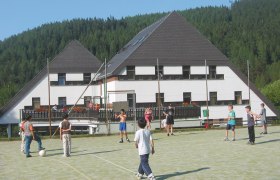 Kinder spielen Fu&szlig;ball auf einem Sportplatz vor einem Hotel in einer bewaldeten Gegend.
