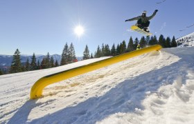 Snowboarder springt über ein gelbes Hindernis im Snowpark Gemeindealpe bei Sonnenschein.