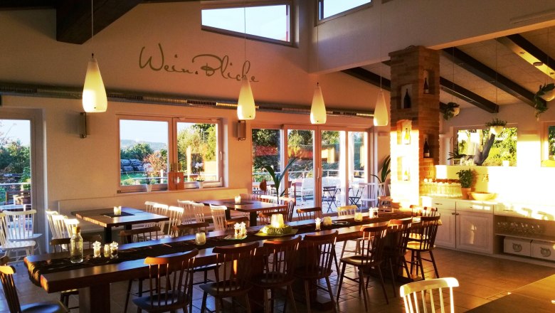 Ein gem&uuml;tliches Gastzimmer mit Holztischen und St&uuml;hlen, dekoriert mit Kerzen und Pflanzen, beleuchtet von warmem Sonnenlicht.