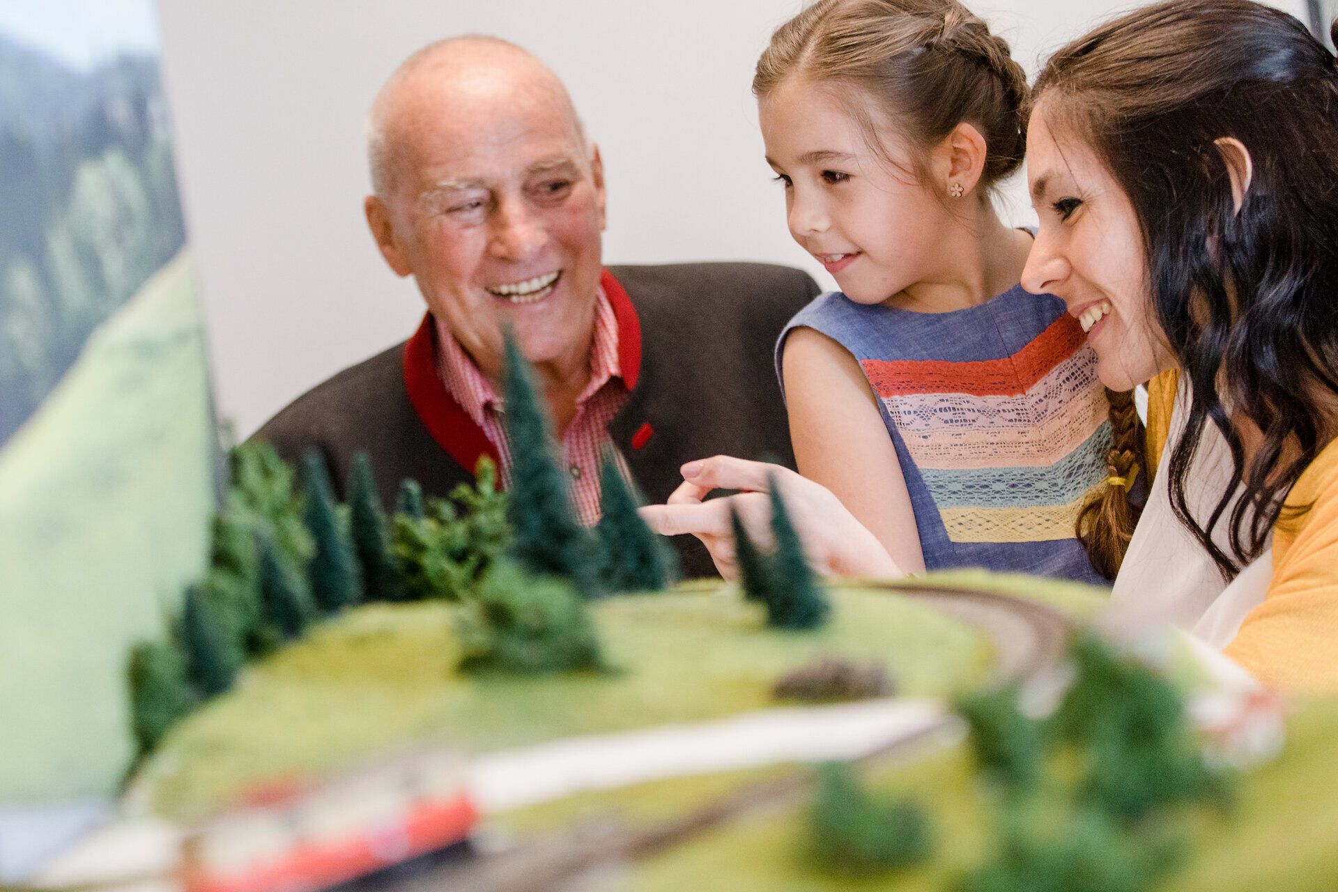 Ein fröhliches Zusammenspiel von Generationen entfaltet sich in diesem Modellbahnmuseum, wo ein Großvater, seine Enkelin und eine Mutter gemeinsam die faszinierenden Züge bewundern. Die leuchtenden Farben der Modelle und die detailreiche Landschaft ziehen die Besucher in ihren Bann und wecken Erinnerungen an unbeschwerte Kindheitstage.