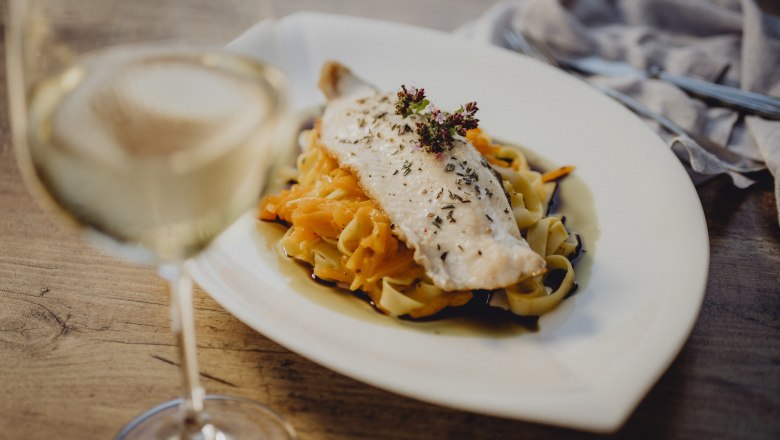 A plate of fish fillet on pasta and vegetables, with a glass of white wine next to it.