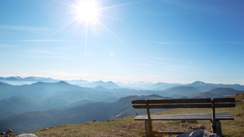 Holzbank auf einem Berg mit Blick auf eine Berglandschaft und die Sonne am Himmel.