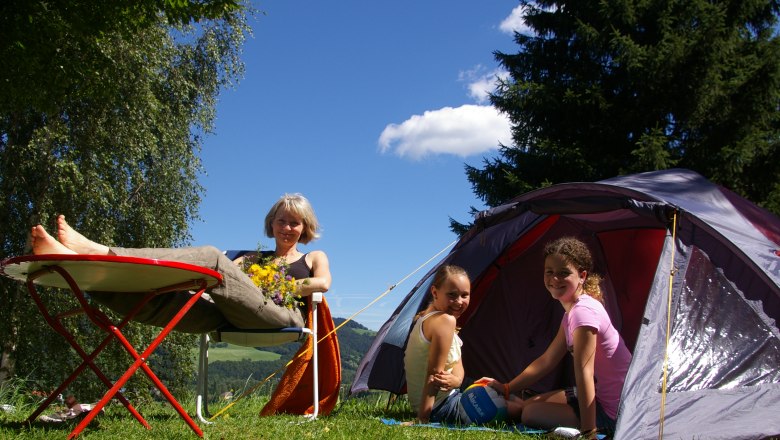 Eine Frau entspannt sich auf einem Campingstuhl neben einem Zelt, während zwei Kinder im Zelt sitzen. Im Hintergrund sind Bäume und blauer Himmel zu sehen.