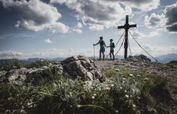 Zwei Wanderer genießen die frische Bergluft und die atemberaubende Aussicht, während sie sich dem Gipfelkreuz nähern. Umgeben von majestätischen Felsen und üppigem Grün, vermittelt die Szenerie ein Gefühl von Freiheit und Abenteuer. Der Sommer in den Bergen ist ein wahres Paradies für Naturliebhaber.