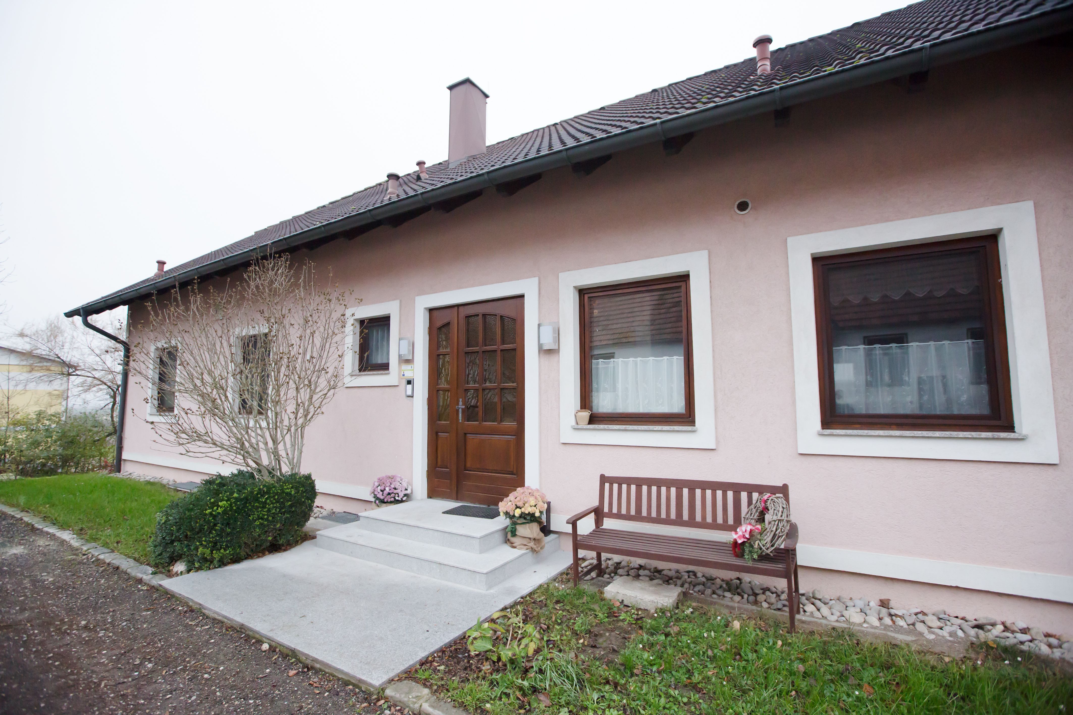 Eingang einer rosa gestrichenen Ferienwohnung mit Holztür, zwei Fenstern und einer Holzbank daneben.