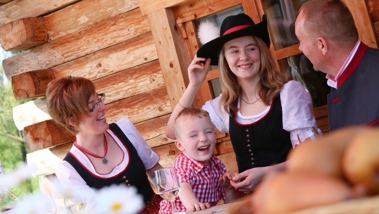 Familie in Tracht vor einer Blockhütte, lachend am Tisch.