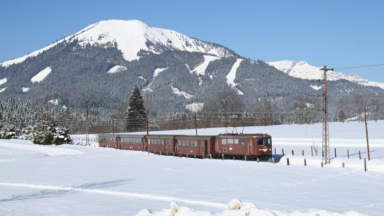 Ein Zug f&auml;hrt durch eine verschneite Landschaft mit einem schneebedeckten Berg im Hintergrund.