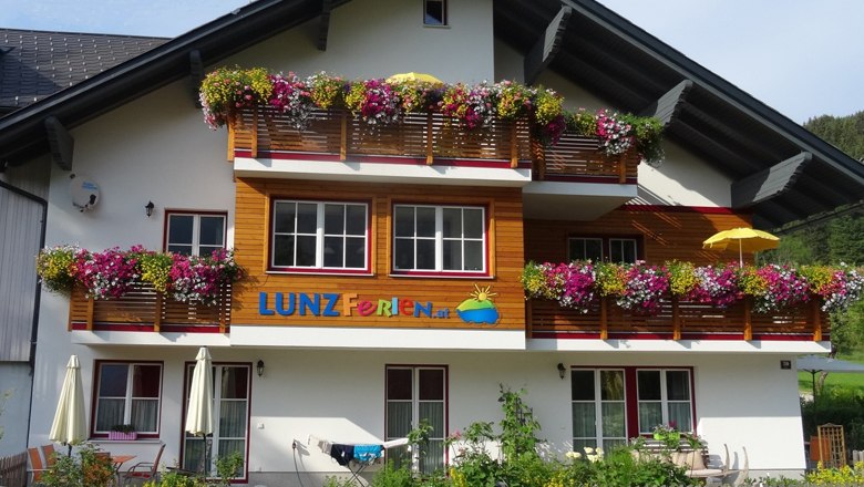 Ein Ferienhaus mit bunten Blumen auf den Balkonen und dem Schriftzug 'LUNZ Ferien'.