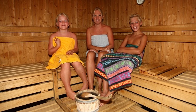 Three people in a sauna, surrounded by wooden walls, clad in towels.