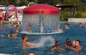 Menschen schwimmen in einem Freibad mit einem großen Wassersprudler in der Mitte.