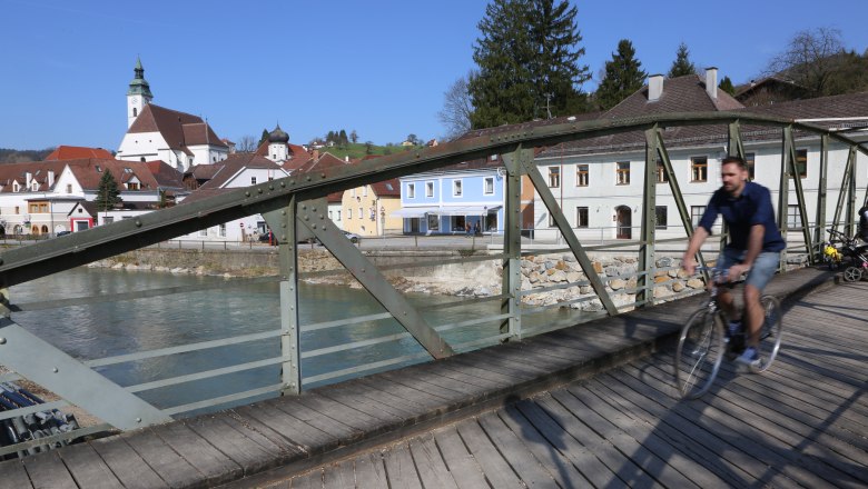 Ein Radfahrer überquert die Töpperbrücke in einer malerischen Stadt mit Kirche und bunten Häusern im Hintergrund.