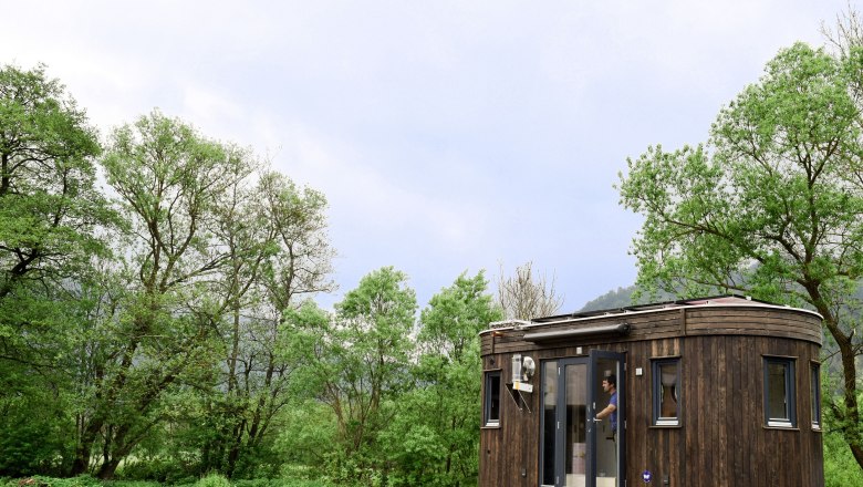 A small, dark caravan with wooden cladding stands in a parking lot in front of green trees and a cloudy sky.