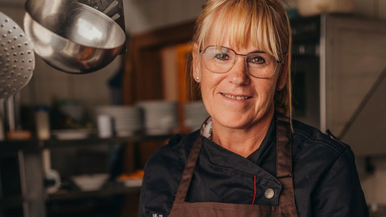 A woman in chef's clothes stands in a kitchen and smiles at the camera.