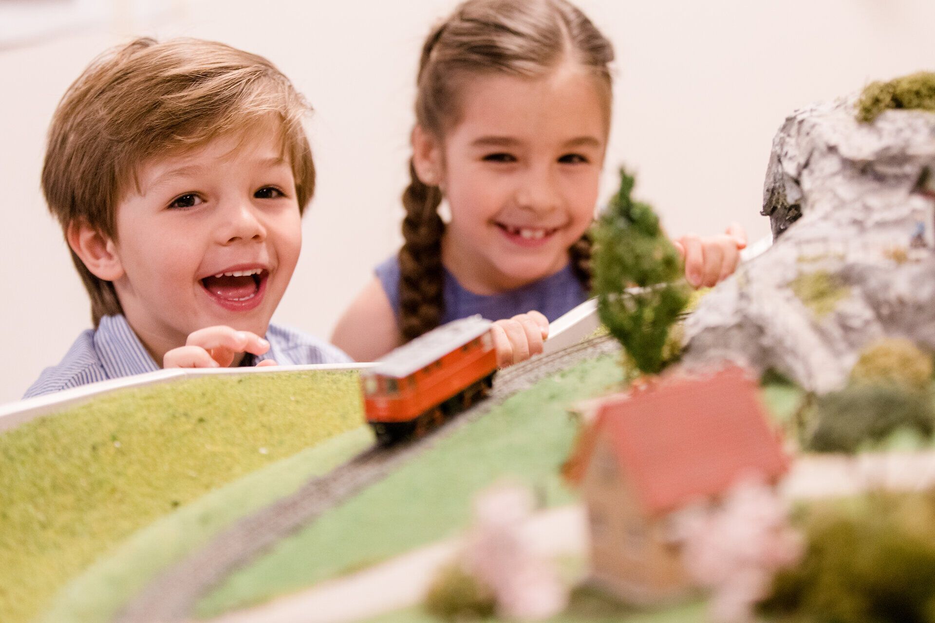 Zwei fröhliche Kinder beobachten gebannt die vorbeifahrende Modelleisenbahn, während sie in eine Welt voller Abenteuer und Fantasie eintauchen. Die detailreiche Landschaft mit kleinen Hütten und Bäumen lässt die Herzen der kleinen Eisenbahnfans höher schlagen. Ein unvergesslicher Moment voller Staunen und Freude.