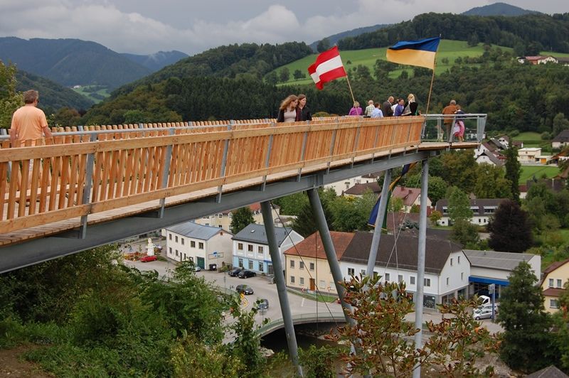 Menschen auf einer Holzplattform mit österreichischer und ukrainischer Flagge, im Hintergrund eine ländliche Landschaft.