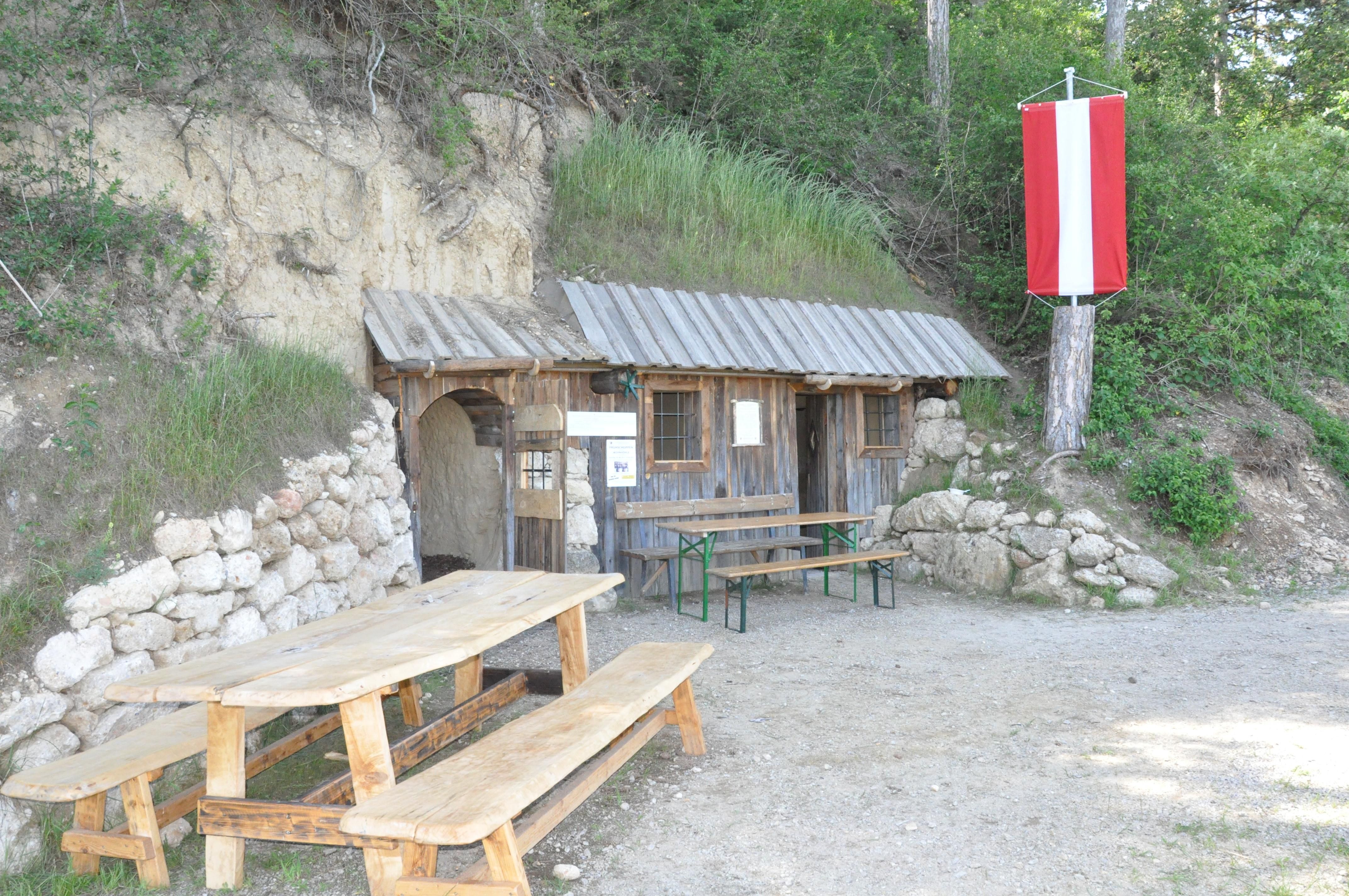 Eine rustikale Holzhütte mit Tisch und Bänken davor, eingebaut in einen Hügel. Eine rot-weiße Fahne hängt an einem Baum.