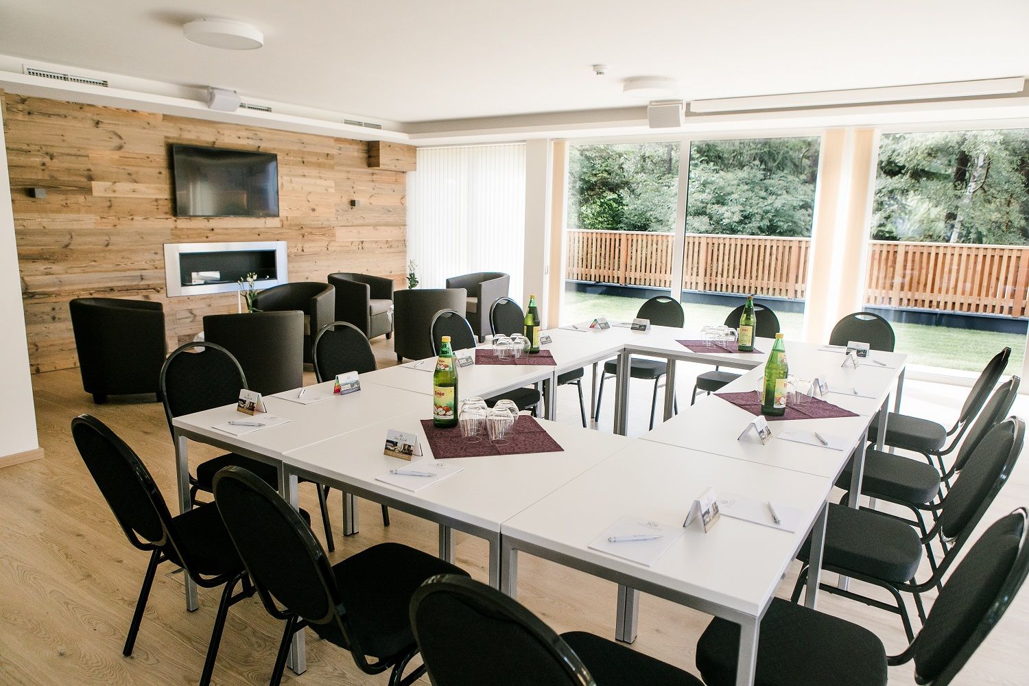 Konferenzraum mit U-förmiger Tischanordnung, Stühlen, Holzwand, Fernseher und großen Fenstern mit Blick ins Grüne.